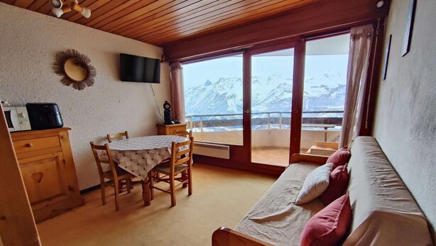 Une pièce chaleureuse avec un plafond en bois, une table à manger avec des chaises, un canapé avec des coussins, un téléviseur fixé au mur et une vue sur des montagnes enneigées, visible à travers de grandes fenêtres.