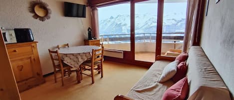 Una habitación acogedora con techo de madera, una mesa de comedor con sillas, un sofá con cojines, un televisor montado en la pared y vistas a montañas nevadas a través de grandes ventanales.
