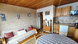 Une pièce chaleureuse avec un plafond en bois, un lit avec une literie à motifs, un canapé avec des coussins, une petite table et un coin cuisine doté de meubles en bois et d’un four à micro-ondes.