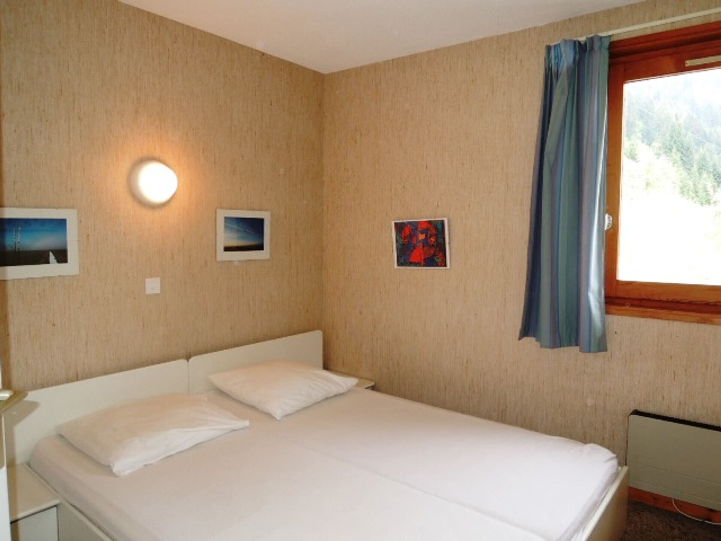 Een eenpersoonskamer met een houten wand, een raam met gordijnen en drie ingelijste foto's.