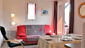 Una habitación acogedora con un sofá rojo, una pequeña mesa lista para comer, un sillón mecededor verde y una ventana con cortinas.