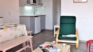 Cocina compacta con microondas, lavamanos y refrigerador. Hay una mesa de comedor con mantel floral y un sillón verde. Una revista y tazas de café están sobre una mesita.