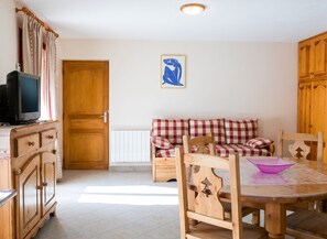 A room with a wooden cabinet, a television, a checkered sofa, a dining table with chairs, and a radiator.