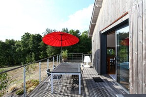 A wooden deck with a table and chairs, a red umbrella, and a glass door leading to an interior space.