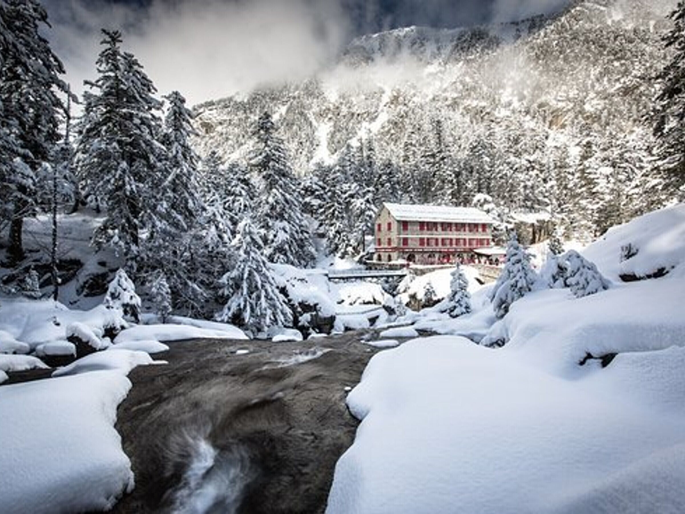 A snow-covered mountain lodge with a large building, surrounded by snow-laden trees and a frozen stream.