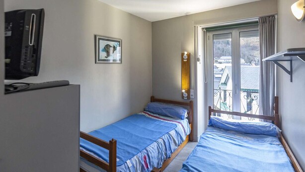 Ein kleiner, kompakter Raum mit zwei Einzelbetten, einem Fernseher, einem gerahmten Bild an der Wand und einem Fenster mit Blick auf Gebäude.