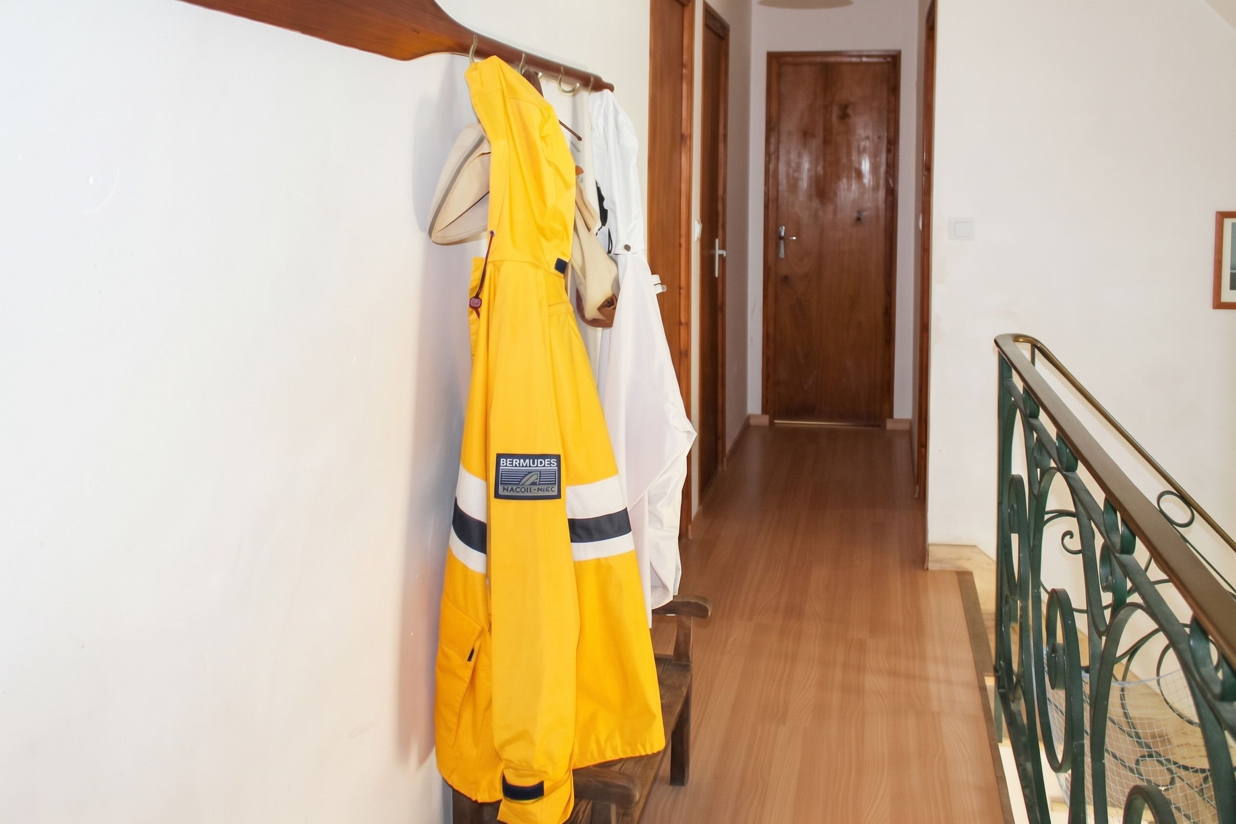 Un couloir doté d’un plancher en bois, un porte-manteau avec des vêtements jaunes et blancs, et une porte en bois au fond.