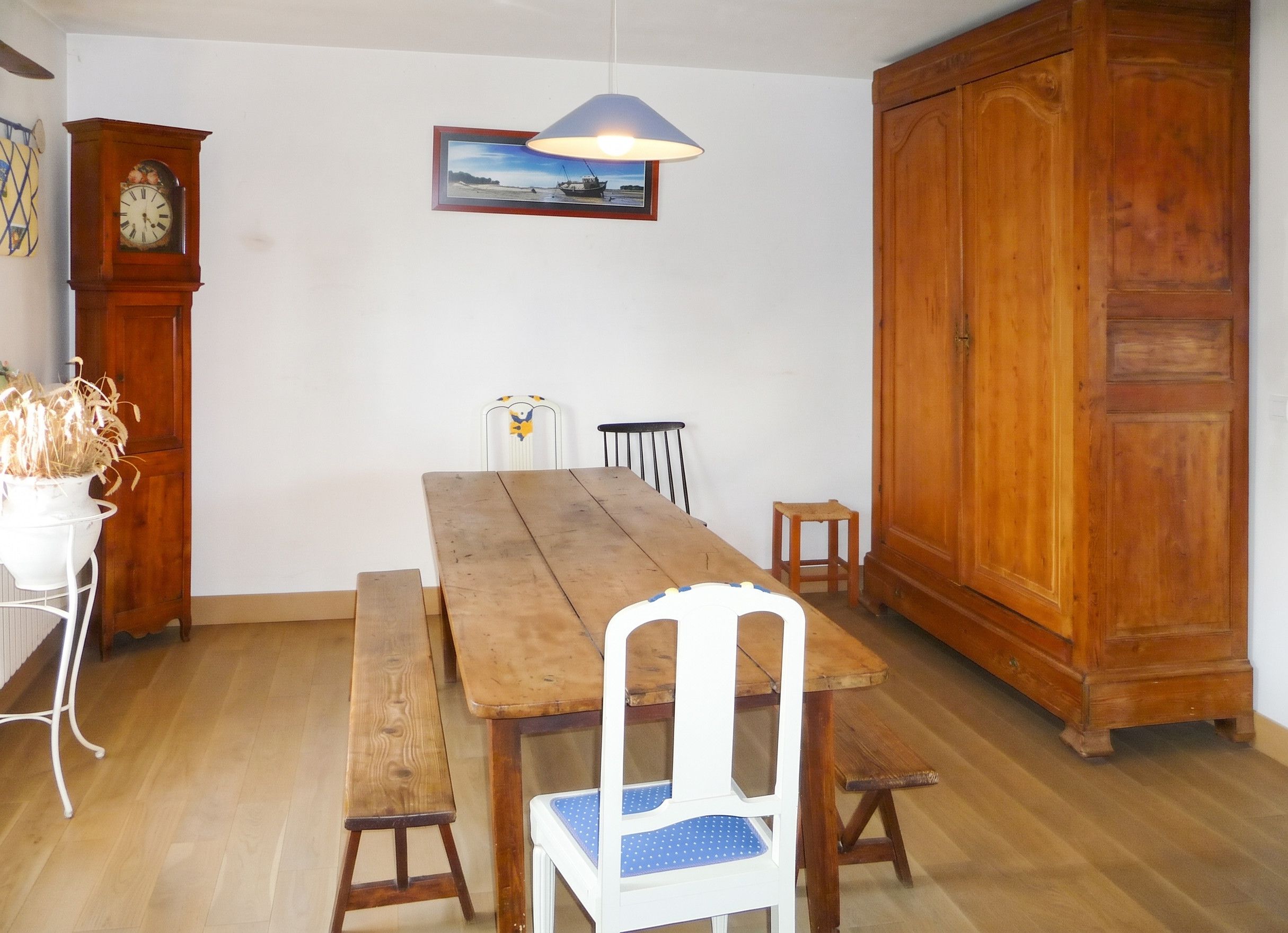 Een eethoek met een houten tafel, bankjes, een witte stoel met een blauwe kussens, een houten kast, een wandklok en een ingelijste foto.