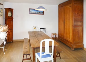 A dining area with a wooden table, benches, a white chair with a blue cushion, a wooden cabinet, a wall clock, and a framed picture. - Homerez - House in Plougrescant. (Plougrescant)