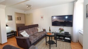 A modern living room with a brown sofa, a coffee table with a bottle and glasses, a flat-screen TV on the wall, and a small black entertainment unit. - lang 80m2 city center          3br (Colmar)