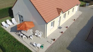 A two-story building with a red-tiled roof, a patio with white plastic chairs and an orange umbrella, and a gravel driveway. - Independent single-storey house near dunes, pets allowed (Les Moitiers-d'Allonne)