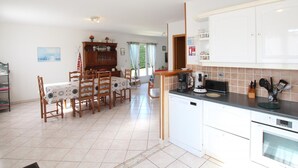 A spacious kitchen with white cabinets, a dining area with a wooden table and chairs, and a view of the outdoors through a window. - Independent single-storey house near dunes, pets allowed (Les Moitiers-d'Allonne)