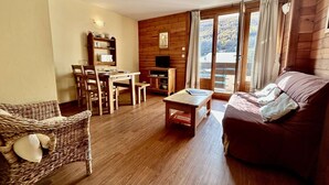 A cozy living room with a wooden floor, a brown sofa, a wicker chair, a dining table with chairs, a small wooden table, and a TV on a wooden wall.