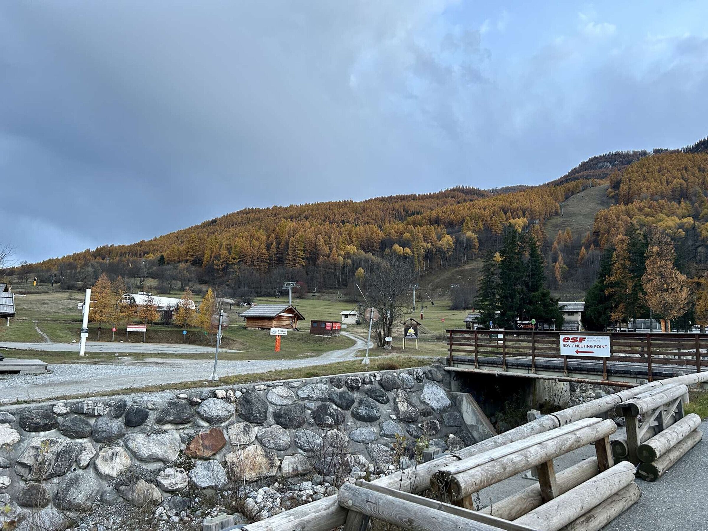 Un chalet de montagne rustique, doté d’un mur en pierre, d’une clôture en bois et d’un panneau indiquant un point de rencontre.