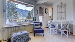 A cozy room with a large window, a dining table with chairs, a blue armchair, and a radiator.