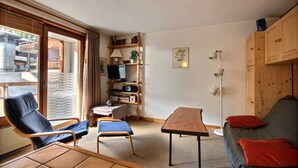Ein gemütliches Wohnzimmer mit einer Couch, zwei Sesseln, einem kleinen Tisch und einem Fernseher. Ein Bücherregal mit verschiedenen Gegenständen und eine Stehlampe sind ebenfalls vorhanden.
