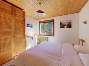 A bedroom with a wooden ceiling, a bed with white bedding, a window with a view of greenery, and a wall-mounted picture.