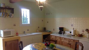 A kitchen with a microwave, sink, and wooden cabinets. There is a window providing natural light and a chandelier hanging above the counter.