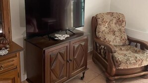 A living room with a flat-screen TV on a wooden cabinet, a floral armchair, and a wall-mounted light fixture.
