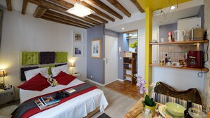 A bedroom with a bed, bedside tables, a nightstand, a bedside lamp, a wall-mounted shelf with items, and a door leading to another room. - D3DG Summer Sweetness (Paris)
