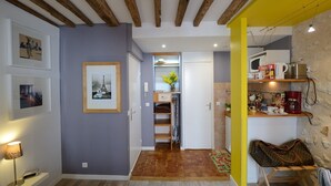 A hallway with a wooden ceiling, a door leading to a kitchen area, and a small seating area with a bag and a lamp. - D3DG Summer Sweetness (Paris)