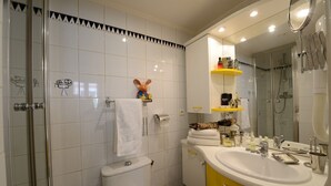 A bathroom with white tiles, a glass shower enclosure, a toilet, a sink with yellow cabinets, and a mirror. - D3DG Summer Sweetness (Paris)