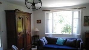 A living room with a dark blue sofa, a wooden cabinet, a patterned armchair, and a window with white curtains. - Villa L'abri Cotier - Sainte-maxime (SAINTE MAXIME)