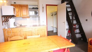 A compact kitchen with wooden cabinets, a microwave, and a toaster. A wooden dining table with chairs is in the foreground. A staircase leads to a lofted area with a red chair. - 2P-Mezzanine Apartment for 6 in Alpe d'Huez (Huez)