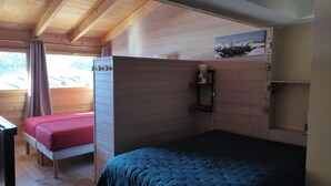 A room with a wooden ceiling, a bed with a blue bedspread, a red bedspread bed, a small shelf with a picture, and a window with curtains. - 2P-Mezzanine Apartment for 6 in Alpe d'Huez (Huez)