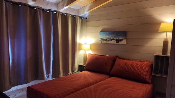 A bed with red bedding, a nightstand with a lamp, a painting on the wall, and a window with curtains. - 2P-Mezzanine Apartment for 6 in Alpe d'Huez (Huez)