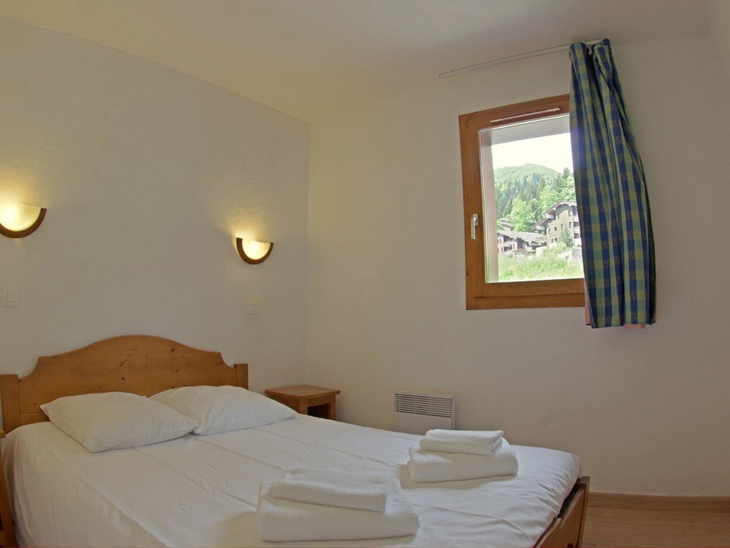 A bedroom with a wooden bed, two bedside tables, a window with a view of buildings, and wall-mounted lighting.