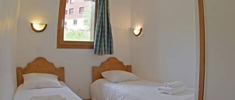 Habitación con dos camas individuales, cada una con cabecero de madera y ropa de cama blanca. Hay apliques de luz a cada lado de una ventana con cortina.