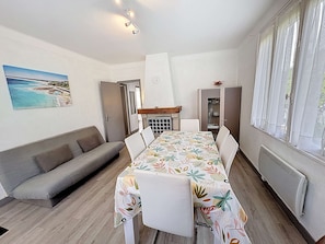 A room with a dining table, white chairs, a grey sofa, a painting of a beach, a cabinet, and a window with curtains.