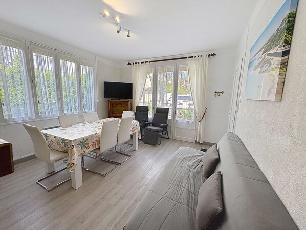Ein Zimmer mit Esstisch, Stühlen, einer Couch, einem Fernseher und großen Fenstern.