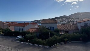 A coastal town with terracotta-roofed buildings, a parking lot, and a view of the sea and distant hills.