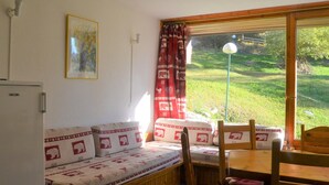 A room with a sofa bed, a dining table, chairs, a lamp, a window with curtains, and a painting on the wall. - Studio for 5, ski-in/ski-out, pets allowed, Arcs 1800 (Les Arcs 1800)