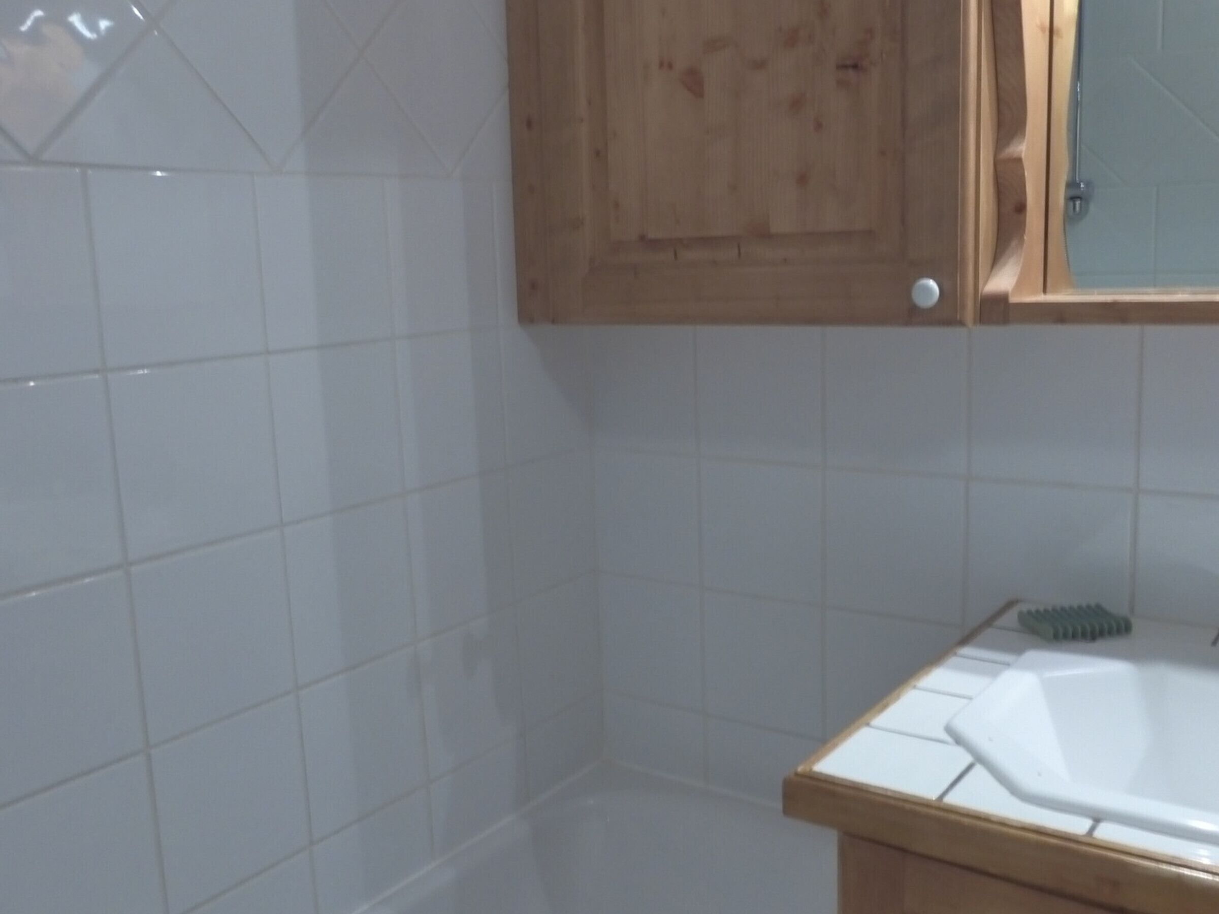 A bathroom with white tiles, a wooden cabinet, and a white sink with a green brush.