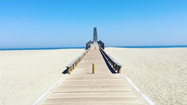 A wooden pier leading to a structure at the end, surrounded by a sandy beach.
