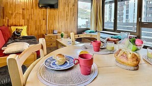 A cozy room with a wooden wall, a TV mounted above a bed, a dining table with breakfast items, and large windows with a view of buildings. - Cozy 2-room apartment for 5 in Tignes, close to slopes and shops (Tignes)