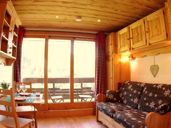 Een gezellig cabin interieur met houten plafond, een bank met bloemenmotieven, een eettafel met stoelen en een groot raam met gordijnen.