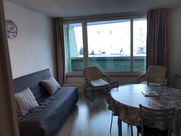 Quarto com sofá, duas cadeiras, mesa e uma janela grande com vista para edifícios.