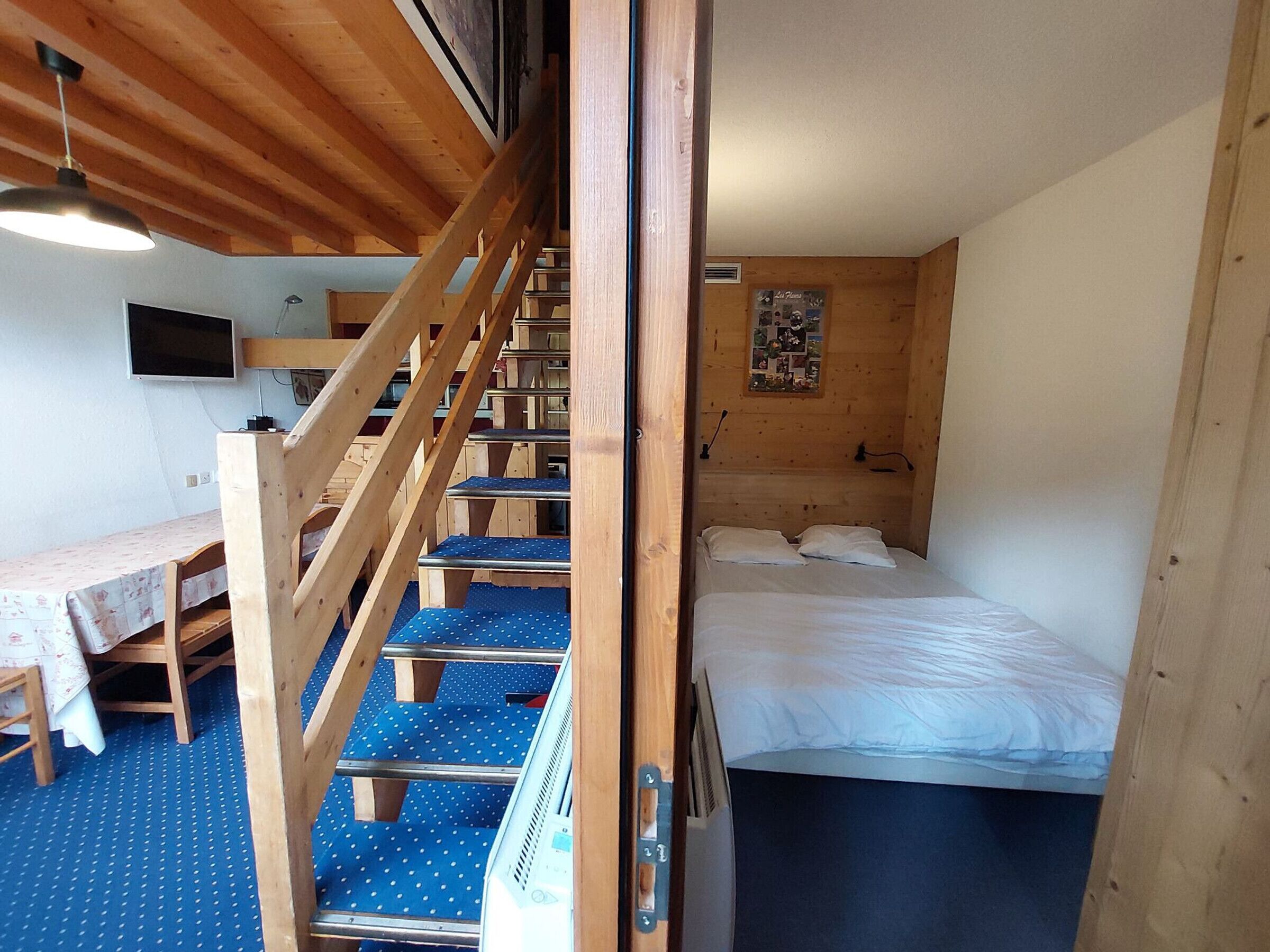 A wooden staircase leading to a bunk bed room with a desk and chair, a television, and a wall with a poster.