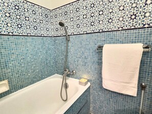 A bathroom with a white bathtub, a showerhead, a towel rack with a white towel, and a candle on the edge of the tub.