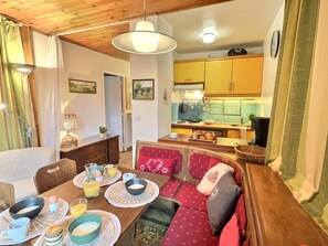 Un comedor acogedor con una mesa de madera, sillas y un banco. Se ve una cocina al fondo con armarios amarillos y una estufa.