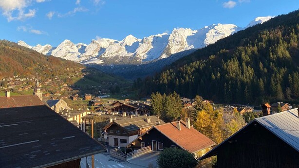 A mountain village with traditional wooden houses, a church with a dome, and snow-capped mountains in the background.