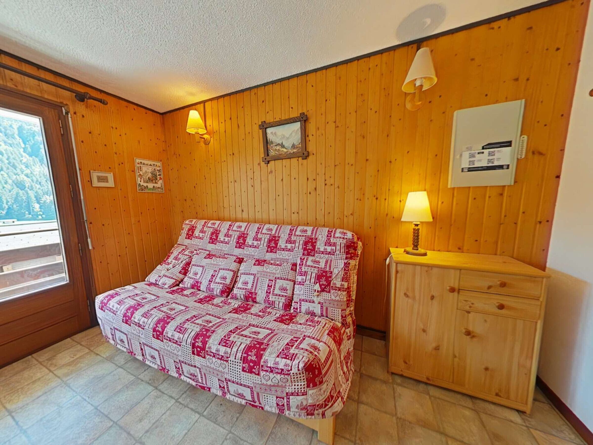 A bedroom with a bed, wooden walls, a wooden cabinet, and a window with a view of trees.