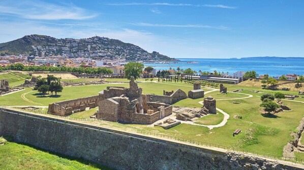 An ancient fortress with ruins, a grassy area, and a view of a coastal city with buildings and a hill in the background.