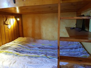 A bunk bed with a patterned bedspread, wooden walls, and a desk with a computer and remote control.