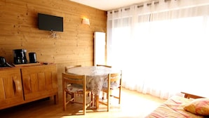 A wooden room with a round table, two chairs, a bed, a TV mounted on the wall, and a window with curtains.