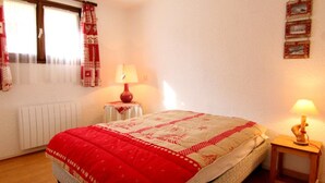 A bedroom with a bed, two bedside lamps, a radiator, a window with curtains, and a wall-mounted picture.